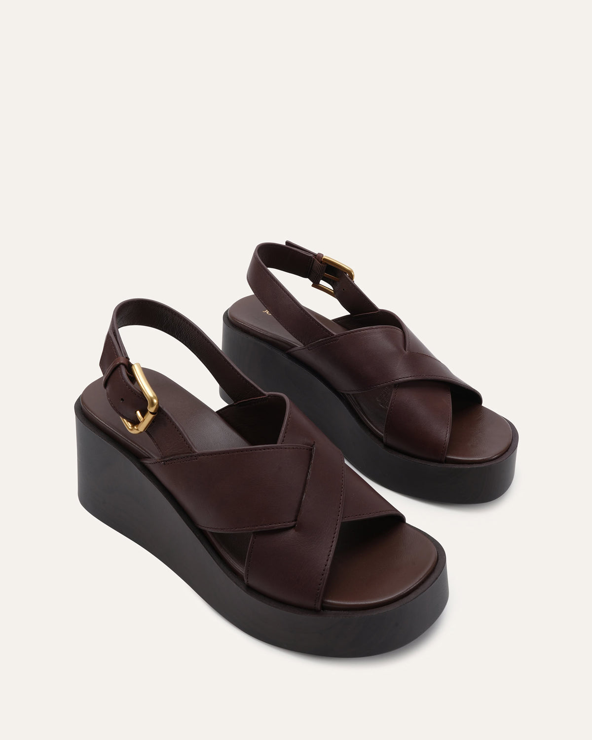 A pair of dark brown leather platform wedge sandals with criss-cross straps and gold-tone buckle details, shown on a light beige background.