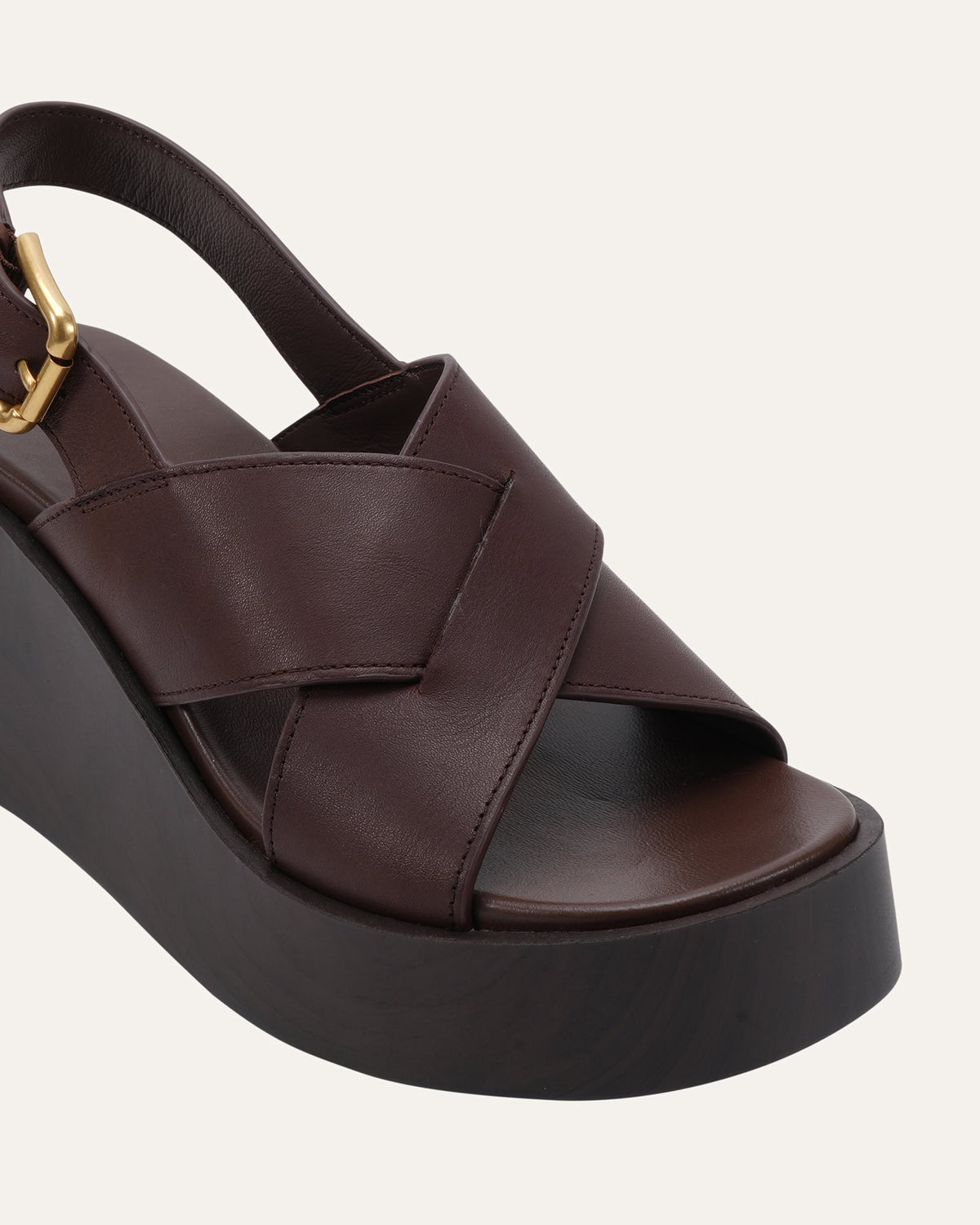 A close-up of a dark brown leather platform sandal with crisscrossing straps, a slingback strap, and a gold-tone buckle, set against a light off-white background.
