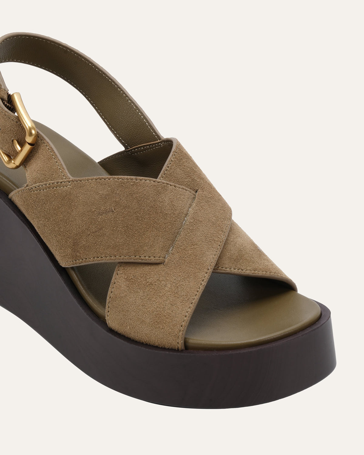 A close-up of a platform wedge sandal featuring taupe suede crisscross straps, a dark brown platform sole, and a gold buckle on the ankle strap, set against a light off-white background.