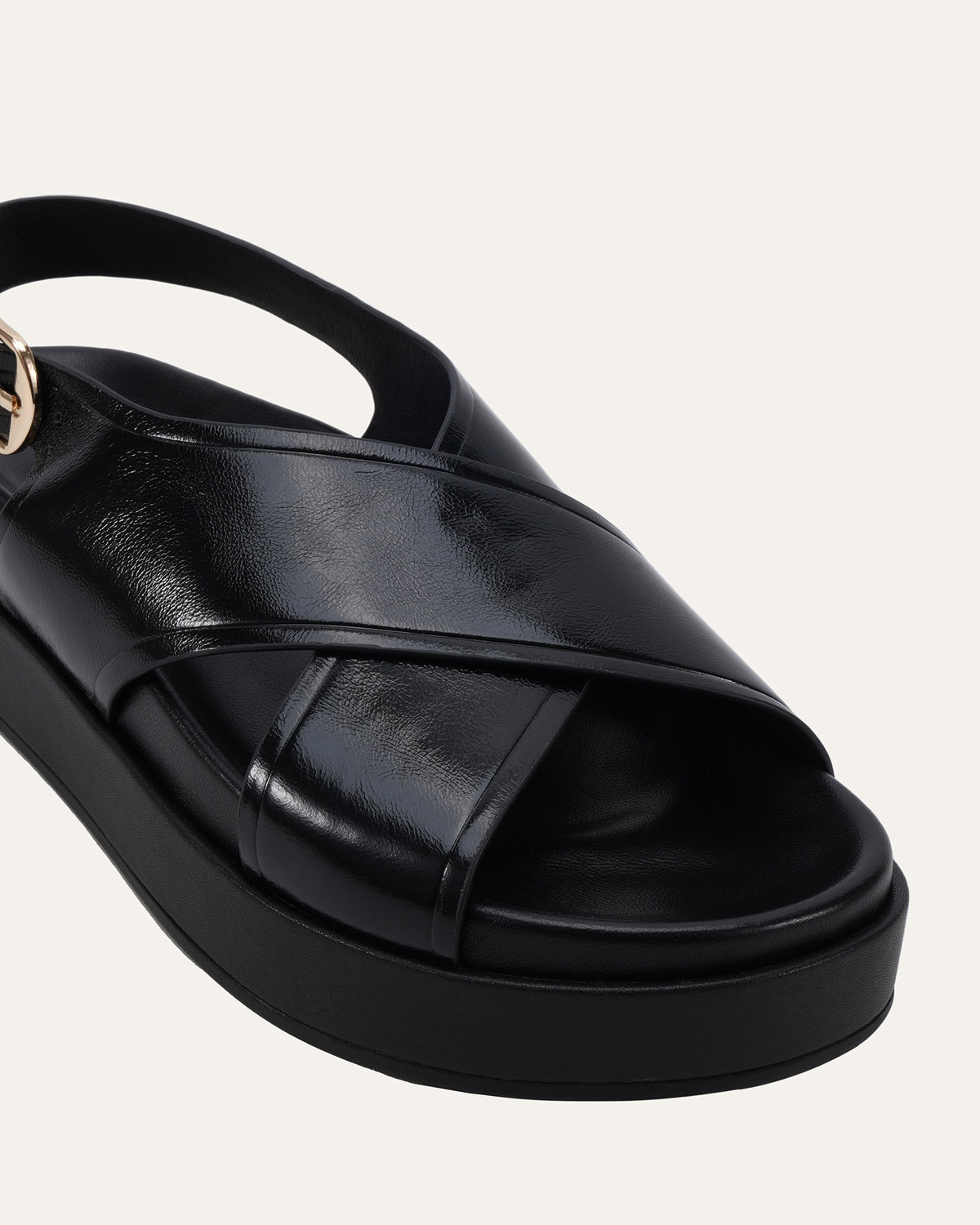 A close-up of a black platform sandal with crisscrossing straps and a slingback strap, featuring a small gold-tone hardware accent on the side, set against a light off-white background.