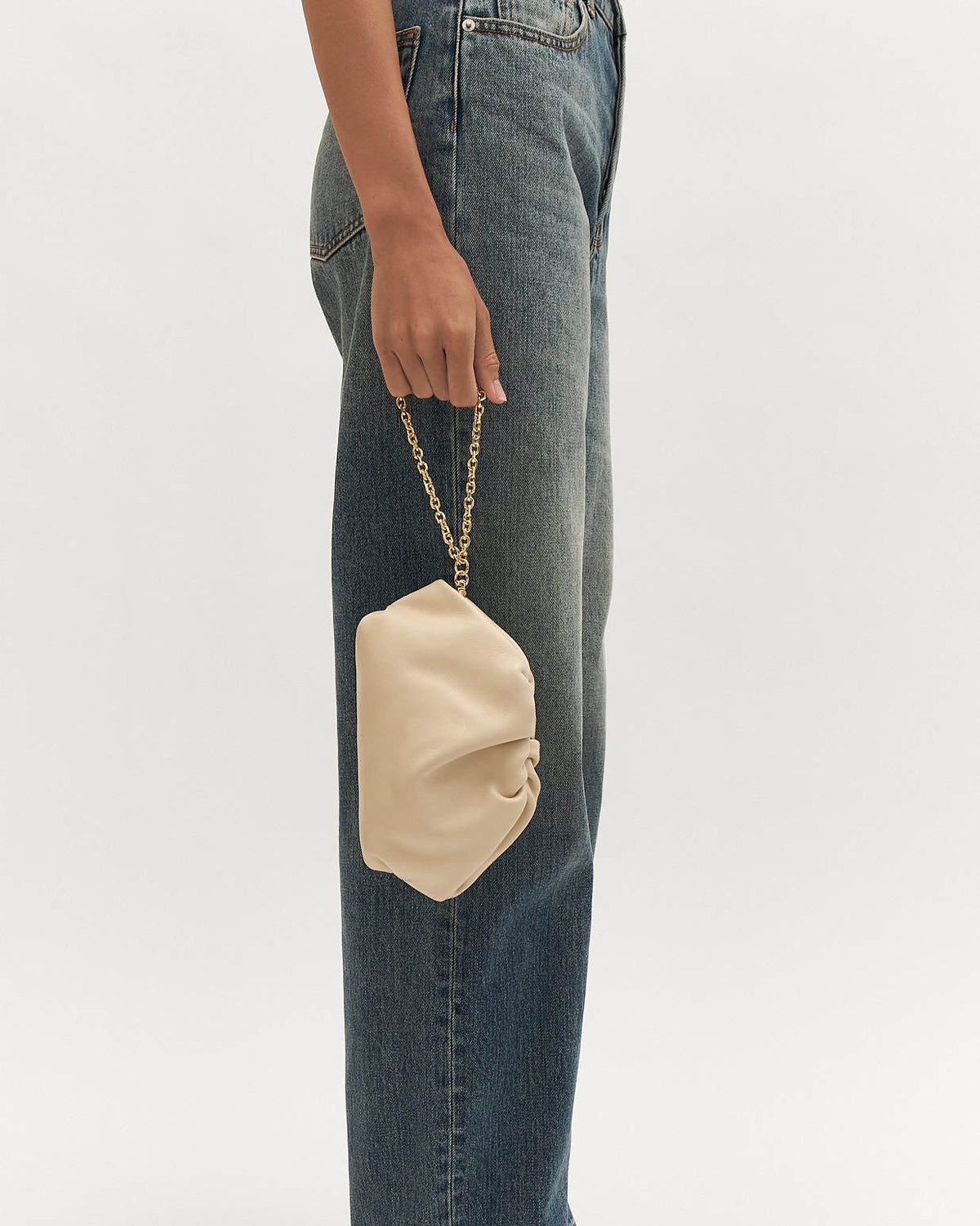 Beige handbag with gold chain held by a person wearing blue jeans on a white background