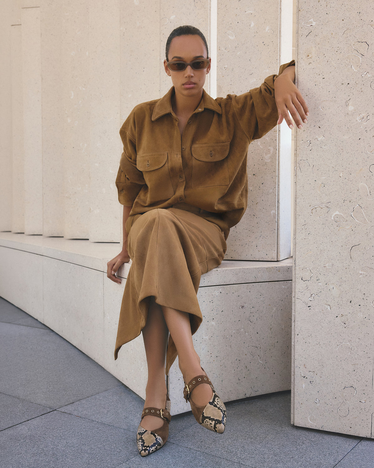 Woman in a brown outfit sitting against a light-colored wall.