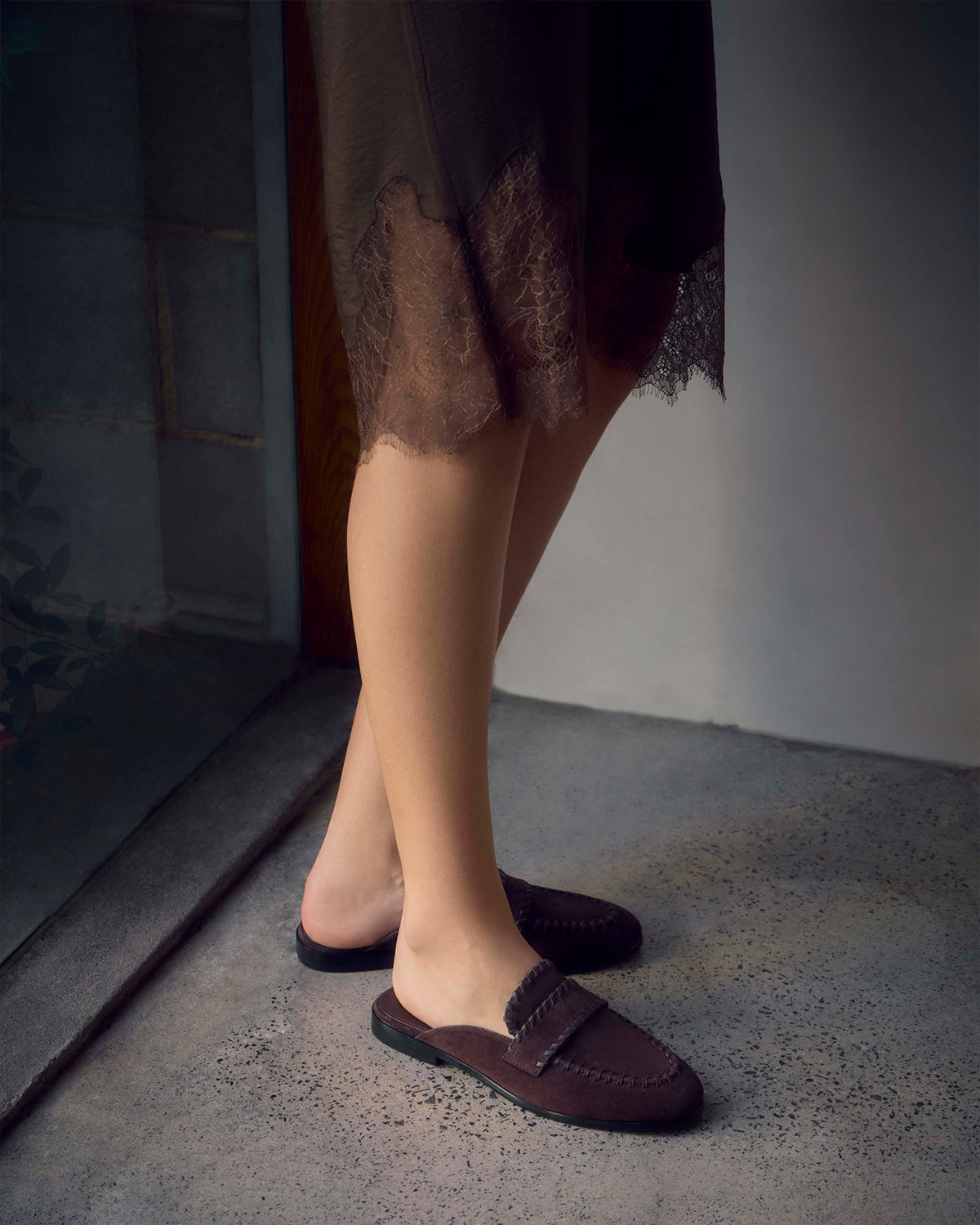 A person wearing dark brown slip-on loafers with a fringed strap detail, standing on a textured concrete floor next to a doorway.
