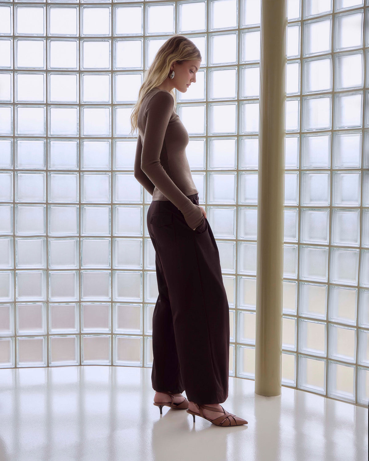 A woman stands in profile in front of a large glass block wall, wearing a fitted taupe long-sleeve top, dark brown wide-leg pants, and nude high-heeled sandals.