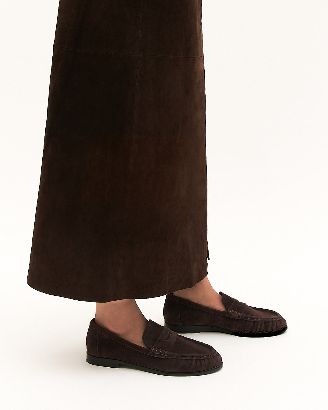 Brown loafers worn with a long brown skirt on a white background