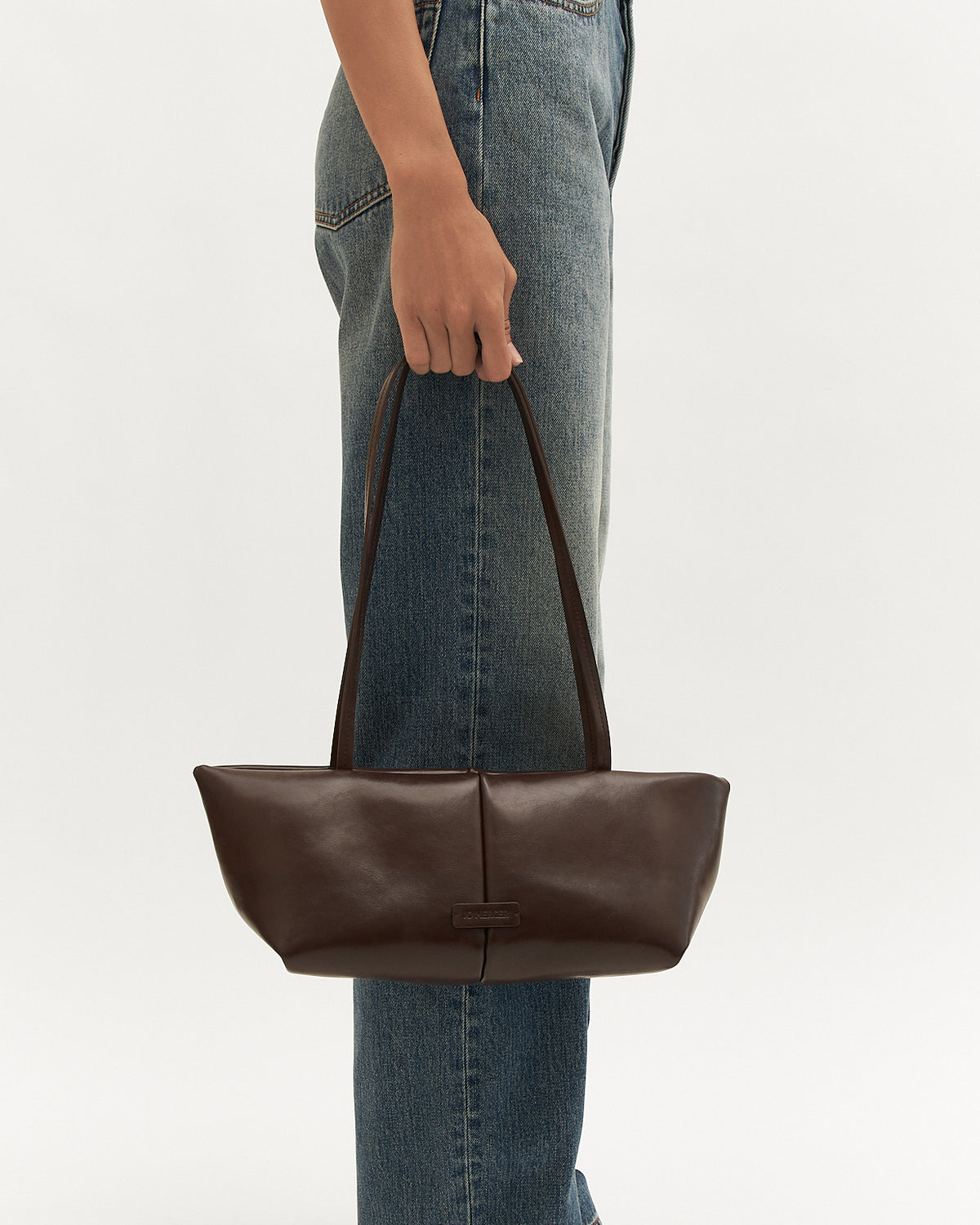 Brown leather tote bag held by a person wearing blue jeans on a light gray background