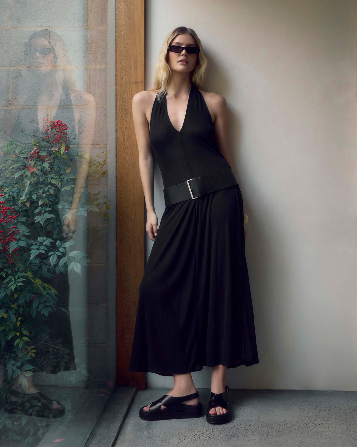 A woman wearing a black halter-neck midi dress with a wide matching belt and black sandals, leaning against a beige wall next to a glass door with a reflection.
