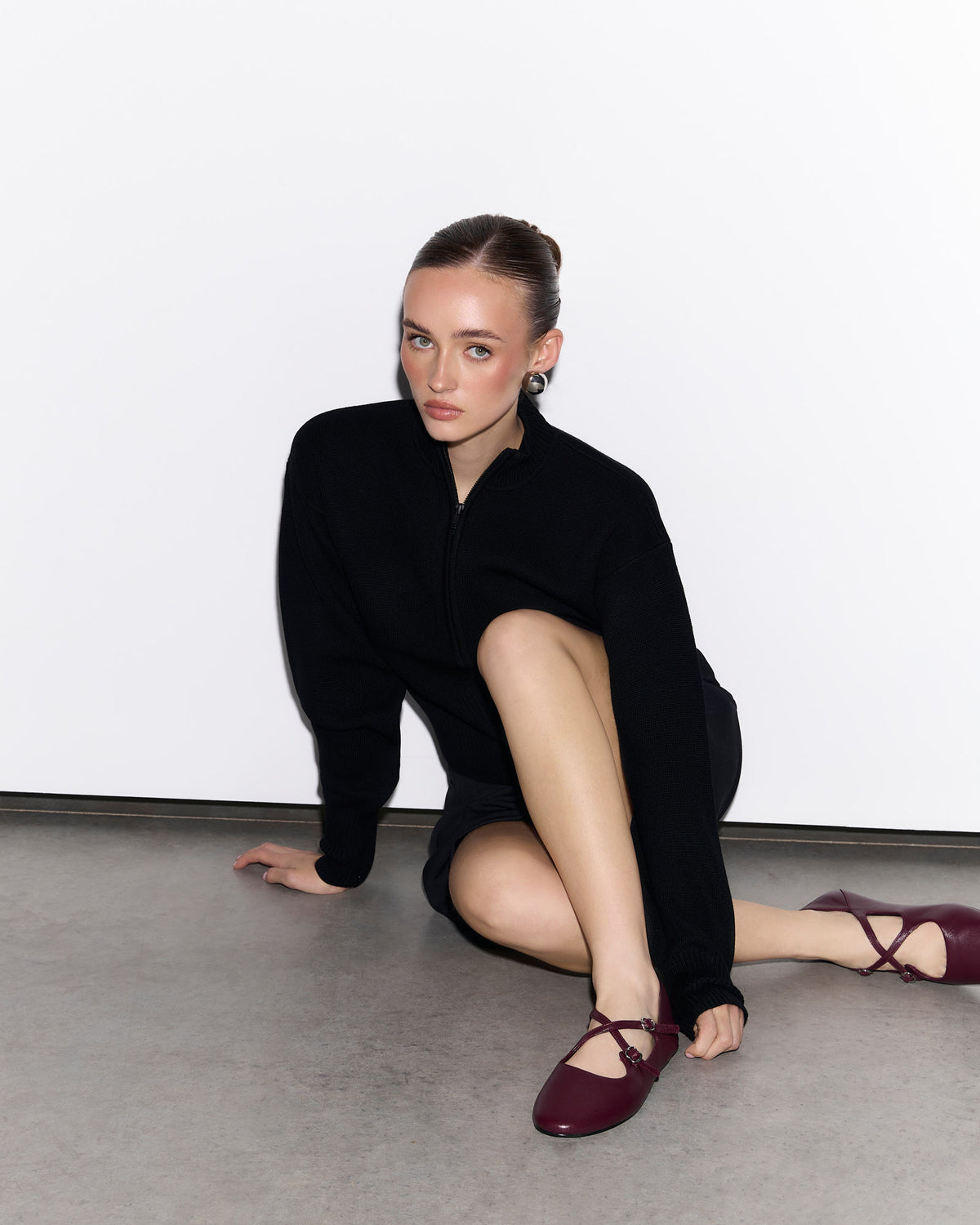 Woman in black outfit with maroon shoes sitting on a gray floor against a white wall